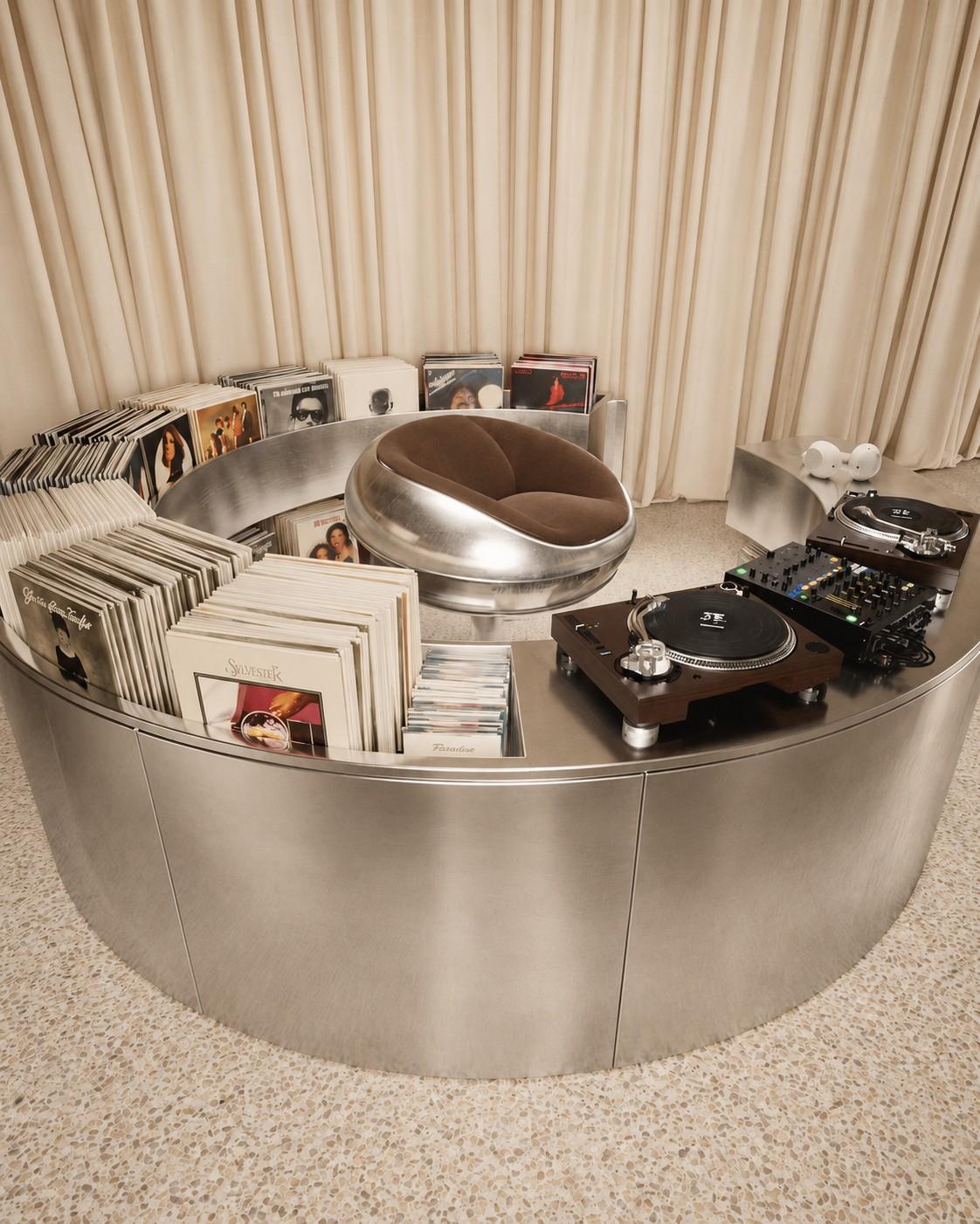 A curved steel DJ booth with vinyl records, turntables and a pod chair
